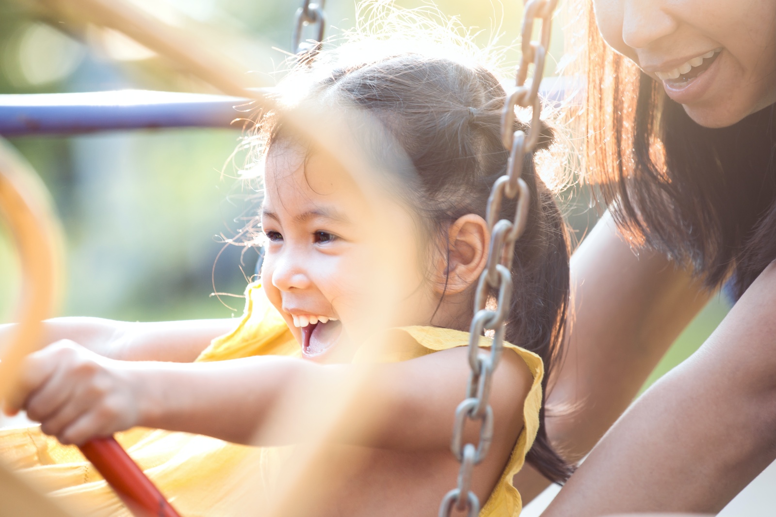 Happy asian little child girl having fun to ride on swings when mother pushing her swings in playground in the sunset time